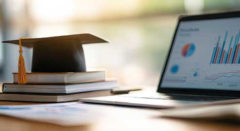 Graduation cap sitting on a stack of books with a laptop beside it.